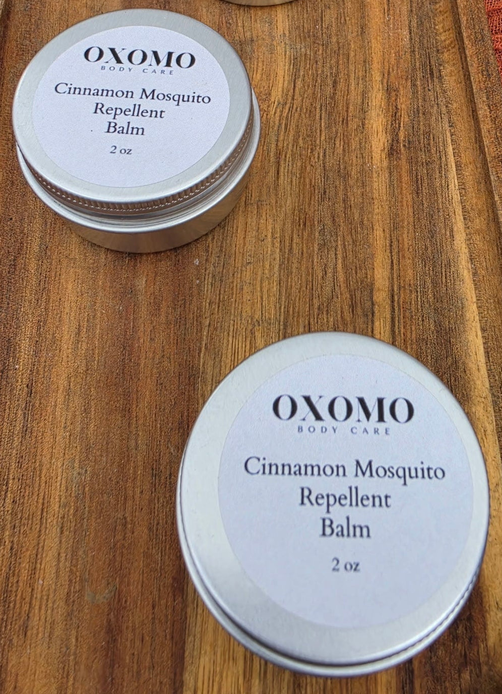 Oxomo cinnamon mosquito repellent balm tins on a wooden tray with an orange fabric background.