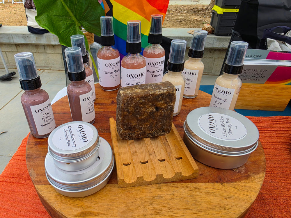 Collection of skincare products including bottles, a bar of soap, and containers on a wooden stand with a rainbow flag in the background.